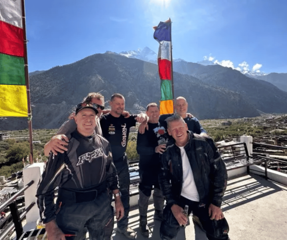 A group of motorcycle adventurers enjoying a break at a scenic rooftop spot with breathtaking mountain views and Tibetan prayer flags fluttering in the wind.