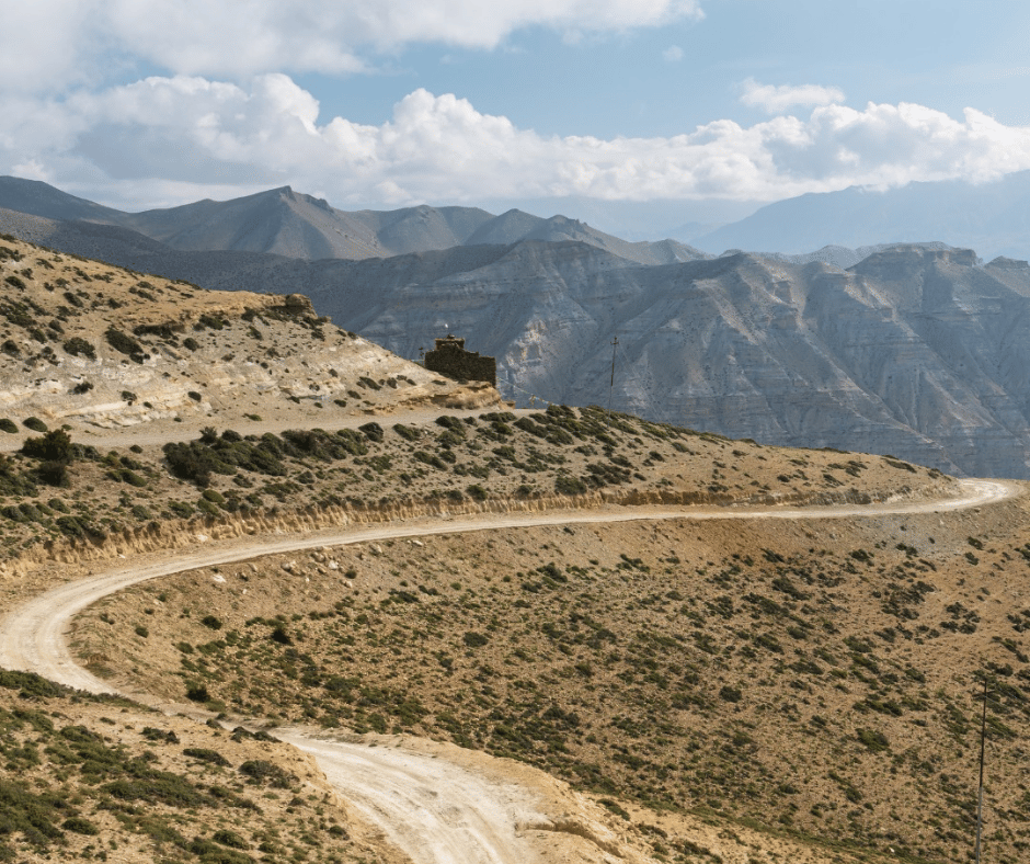 A winding dirt road cutting through the rolling Mustang hills, leading to an ancient, isolated structure perched on the mountainside.
